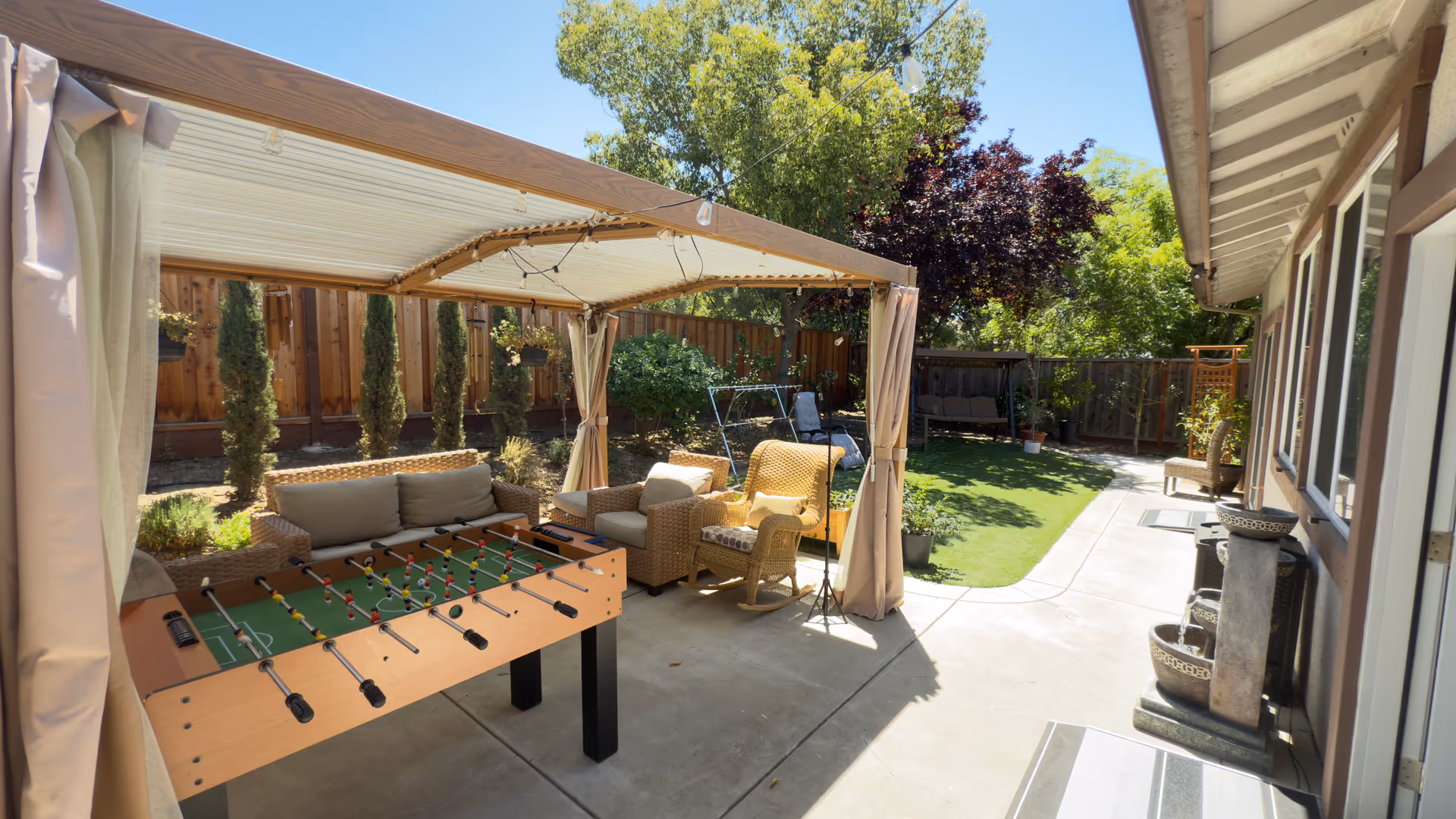 Outdoor patio with a canopy, wicker seating, foosball table, and grassy yard.