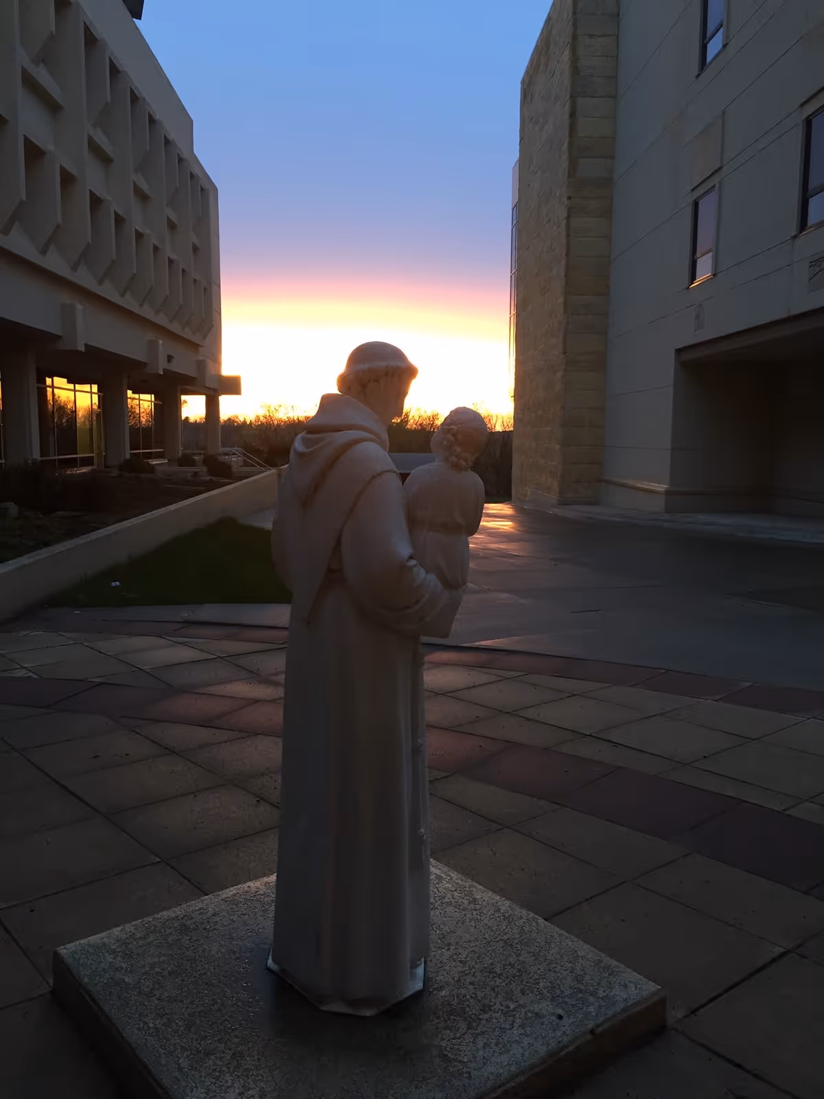 Outdoor statue of a robed saint holding a child near St Anthony Regional Hospital at sunset.