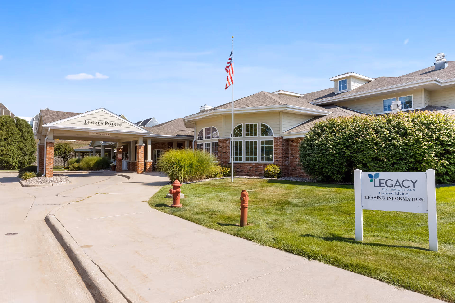 Front entrance and driveway of the Legacy Pointe senior living facility with a canopy, American flag, and a leasing information sign on the lawn.