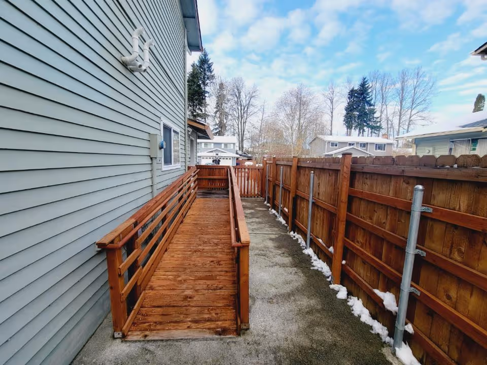 Wooden ramp runs along the side of a light-blue house, fenced yard, snowy ground.