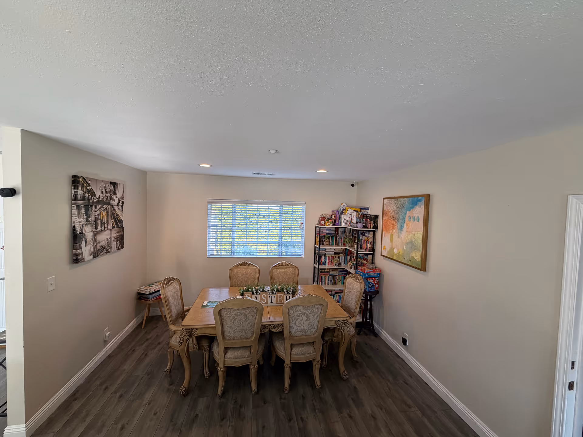 An interior dining room with a wooden table, ornate chairs, a bookshelf, and wall art.
