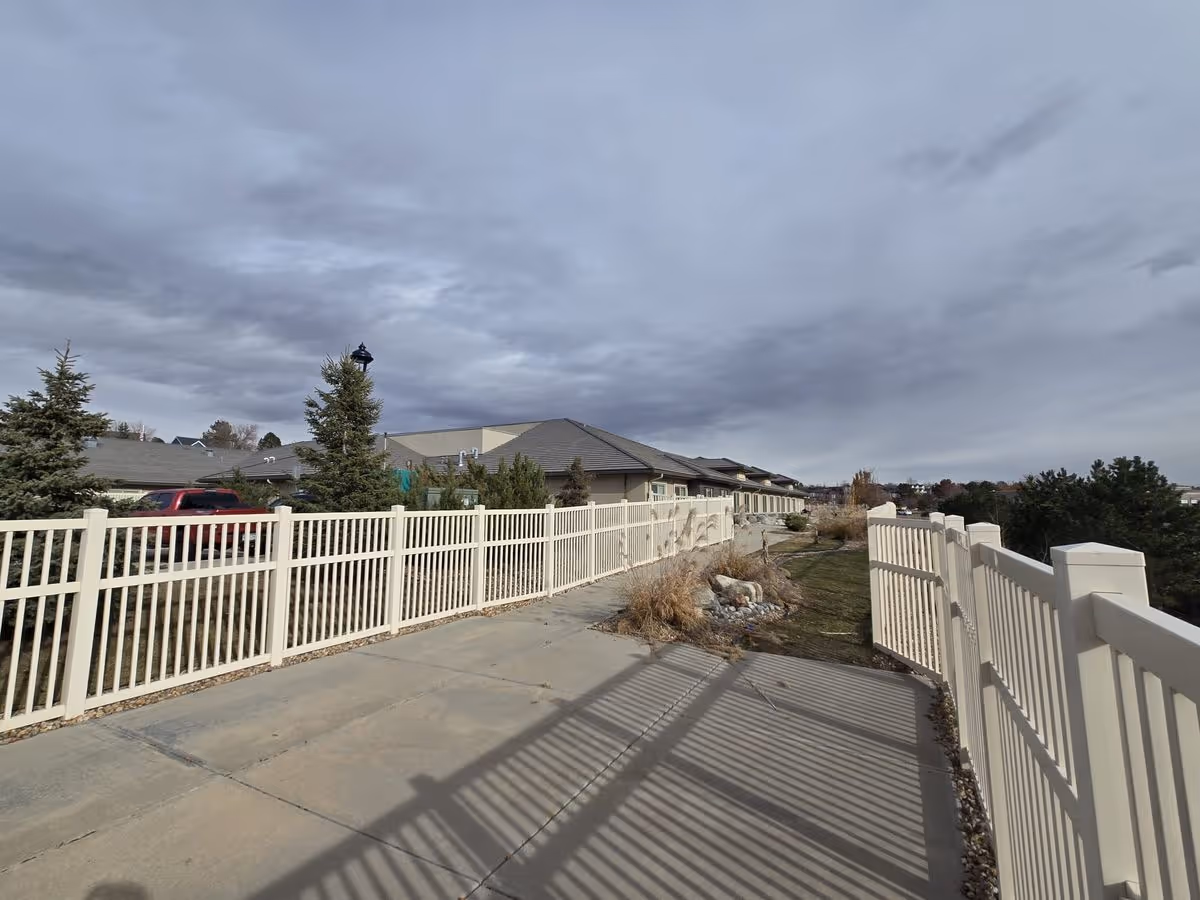 Concrete walkway beside a white fence leading past a single-story memory care building under a cloudy sky.