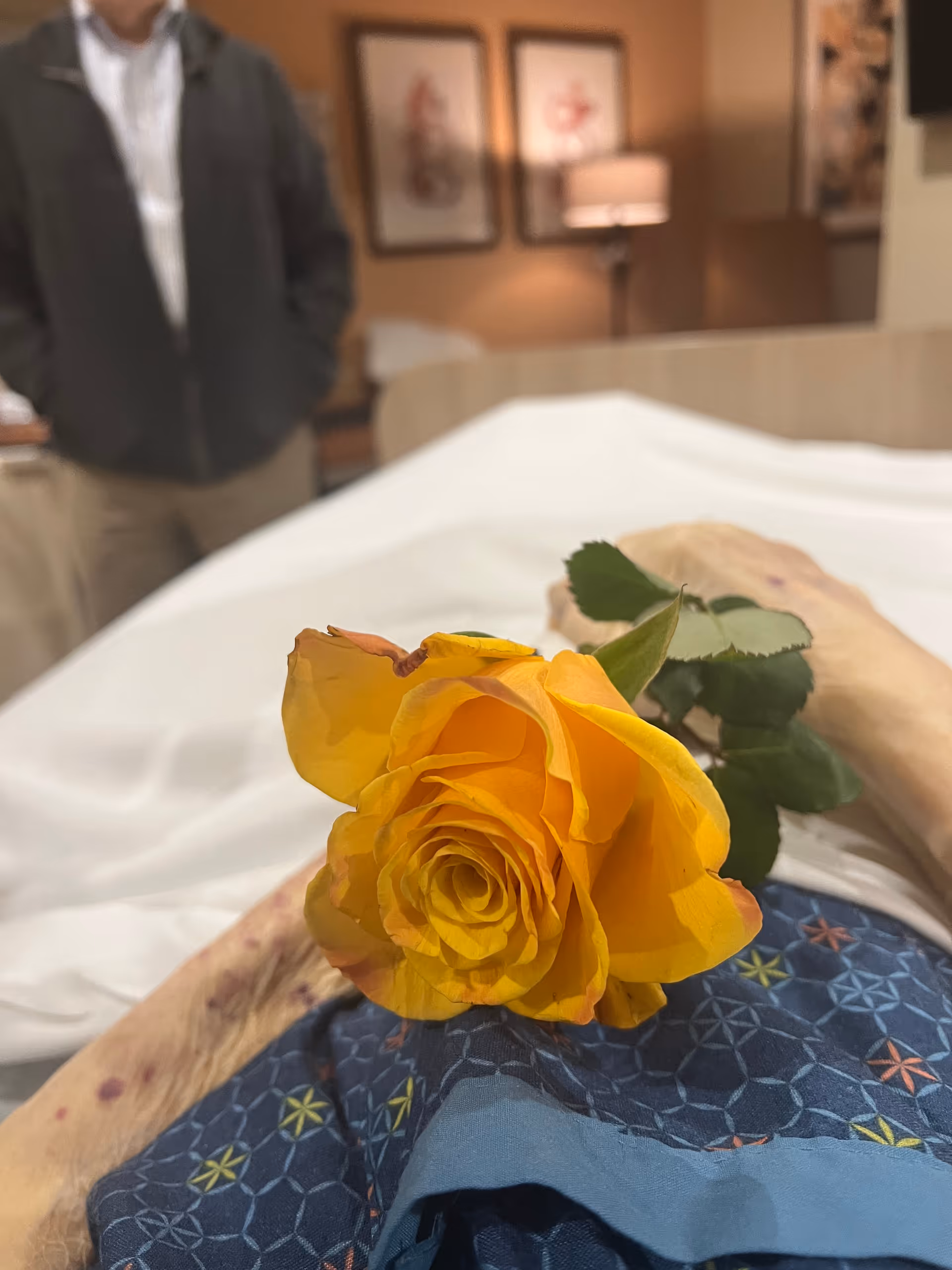 Yellow rose on a patient's bed in hospice room, with a blurred visitor in the background.