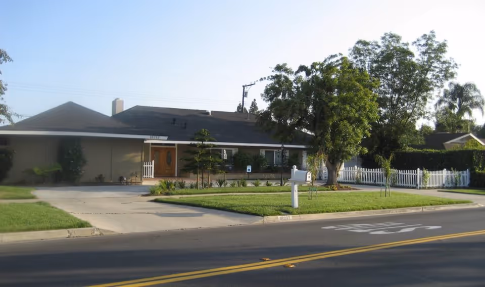 A single-story beige senior living facility with a manicured lawn, front entrance, and white fence.