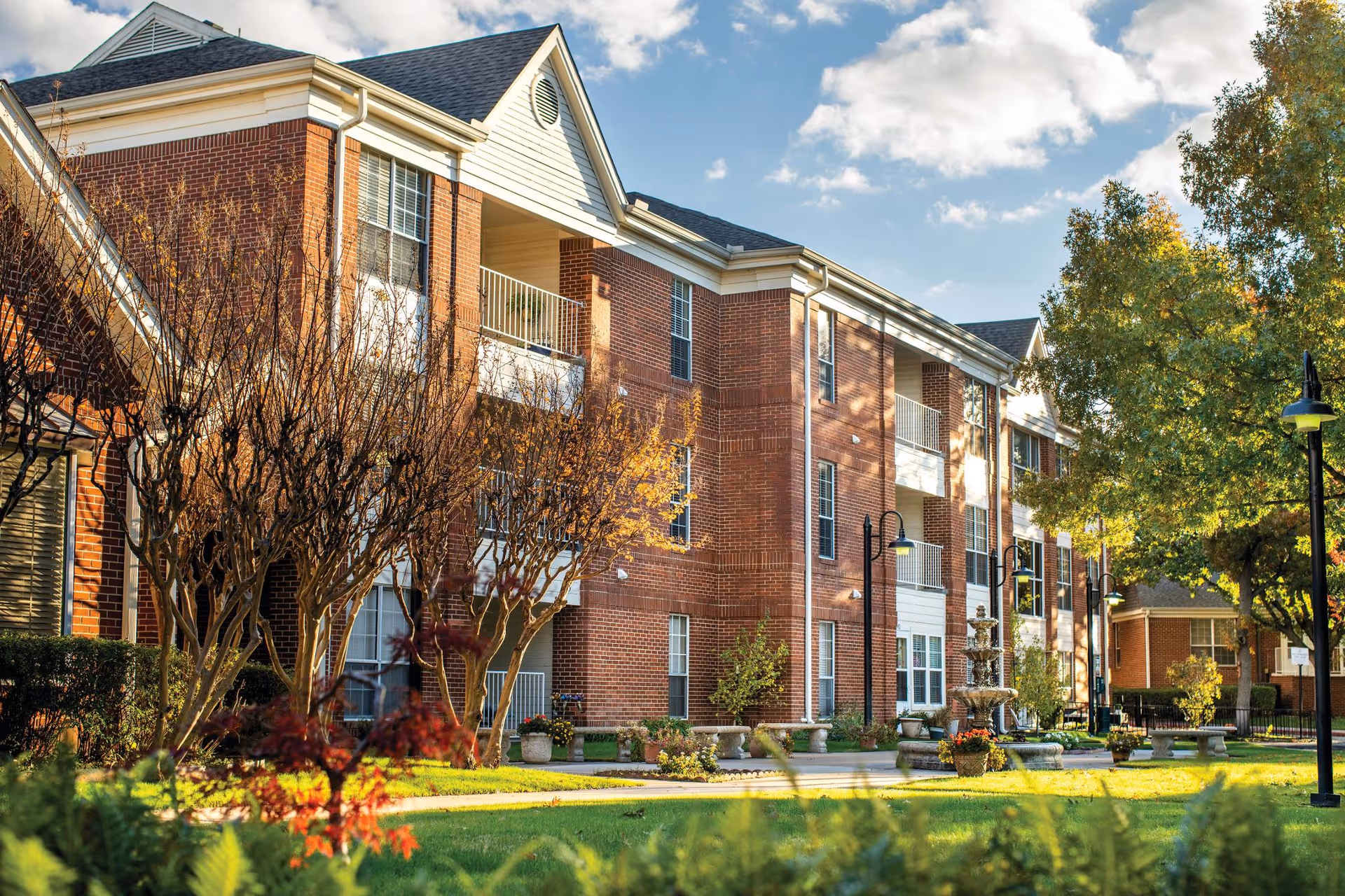 Exterior view of a multi-story brick senior living facility with balconies, surrounded by trees and landscaped greenery under a partly cloudy sky.
