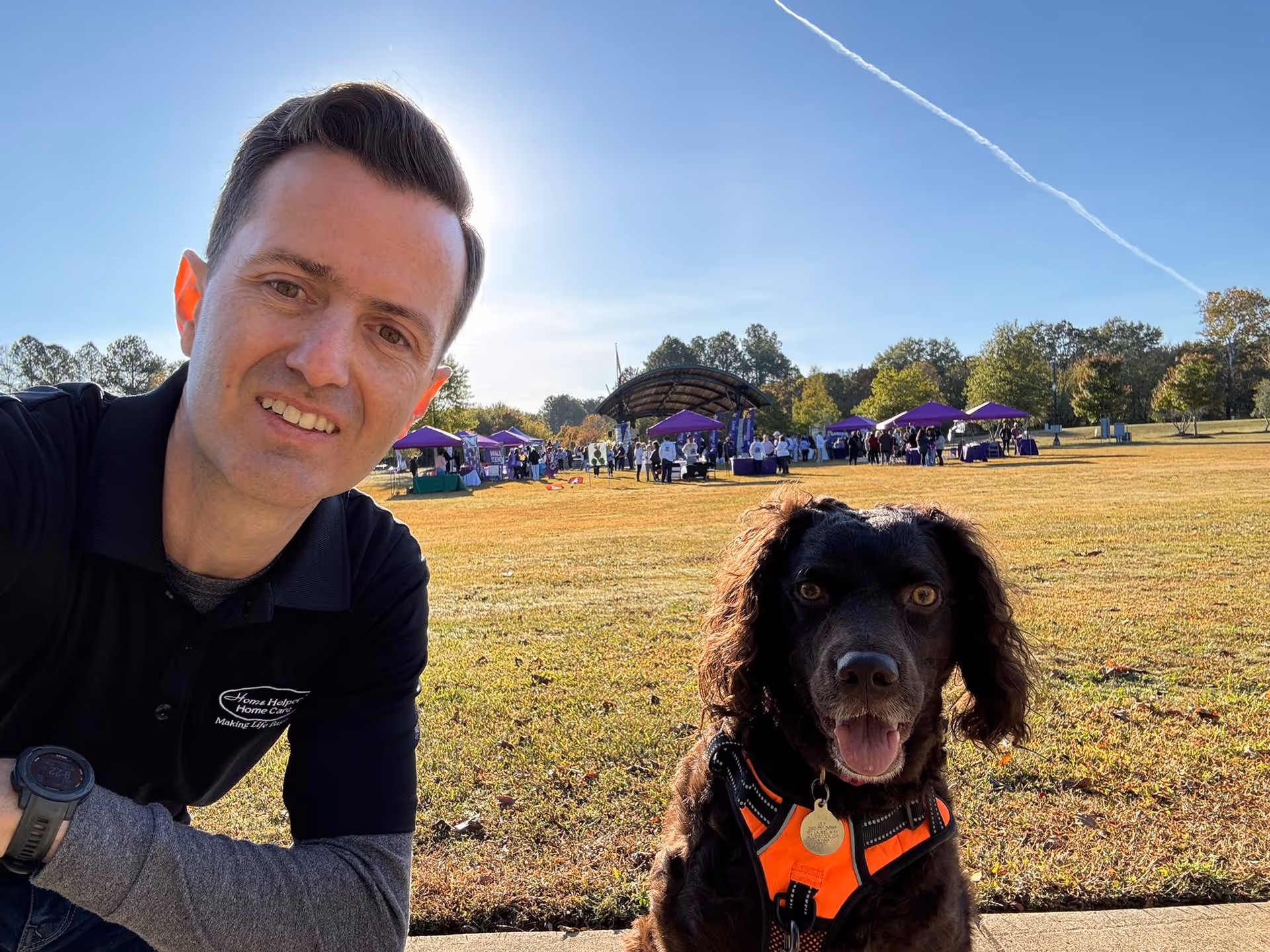 Caregiver in a polo takes a selfie with a dog outdoors at a park event.
