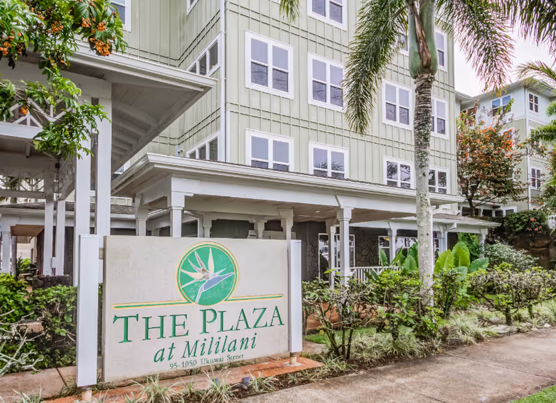 The Plaza at Mililani exterior building with a sign and tropical landscaping.