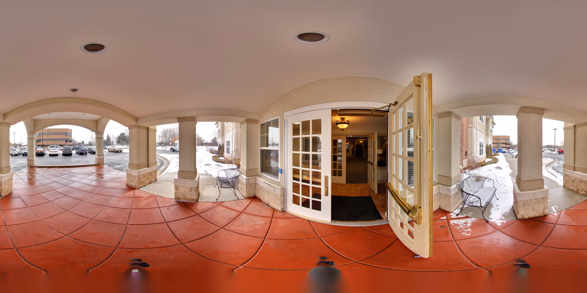 Covered entrance area of a senior living facility with red tiled floor, beige pillars, and open double glass doors leading inside. Outside, there are benches on either side and a parking lot with several cars visible in the background. Snow is on the ground near the building.