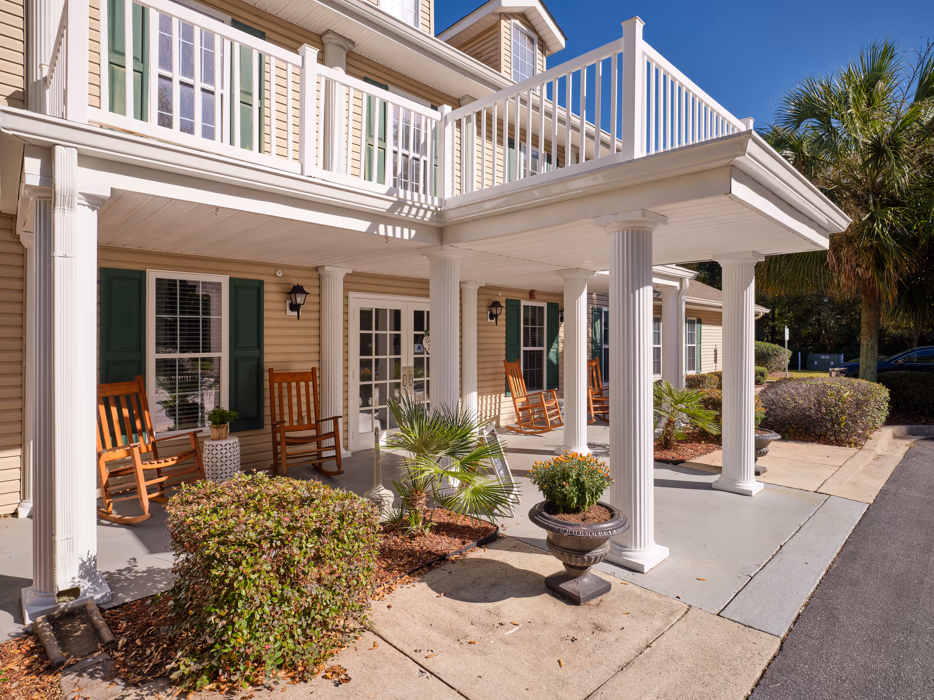 Covered front entrance of a building with white columns, rocking chairs on the porch, and potted plants.