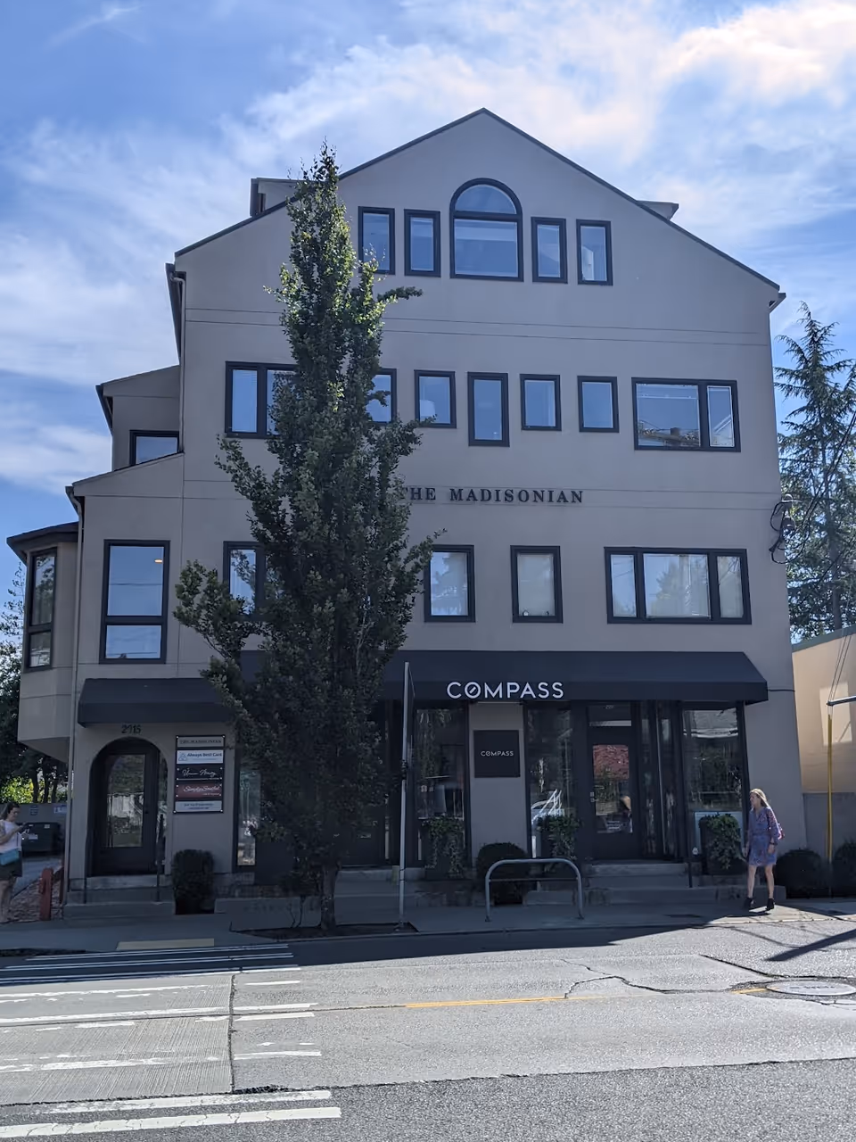 Multi-story beige building with Compass storefront on a city street, tree in front.