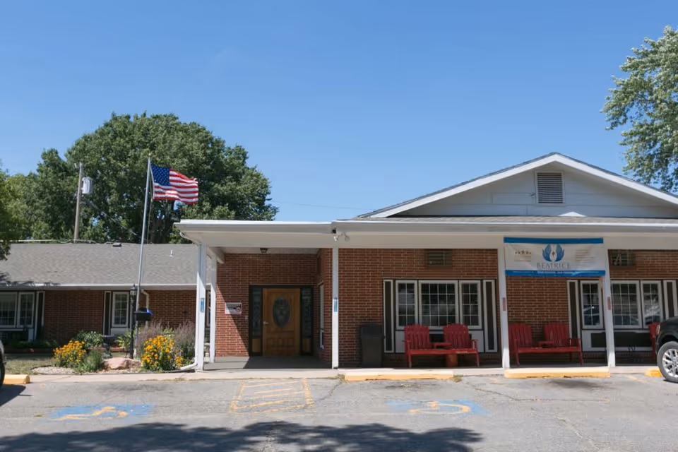 Front exterior view of Beatrice Health and Rehabilitation building with a brick facade, an American flag on a flagpole, red benches on the porch, and a clear blue sky.