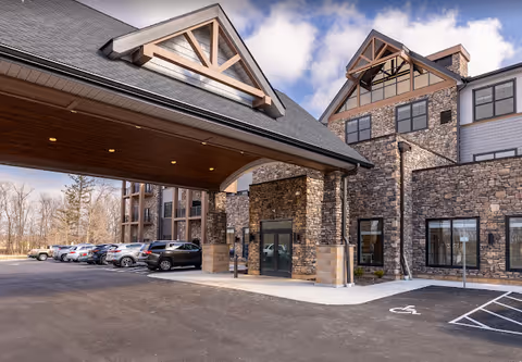 Entrance of a stone-clad senior living facility with a covered porte-cochère, parked cars, and a multi-story building under a partly cloudy sky.
