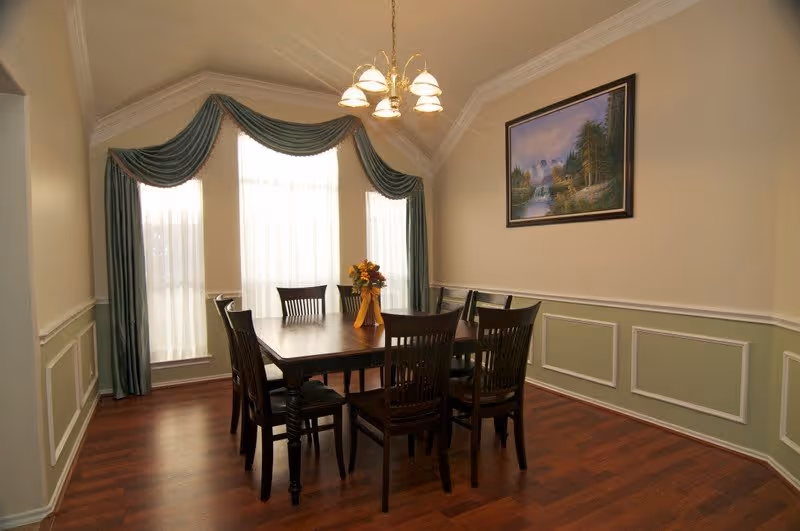 A dining room with a dark wooden table and six matching chairs on a polished wooden floor. The room has three tall windows with sheer white curtains and green drapes with valances. A chandelier with five lights hangs above the table, and a framed landscape painting is on the wall.