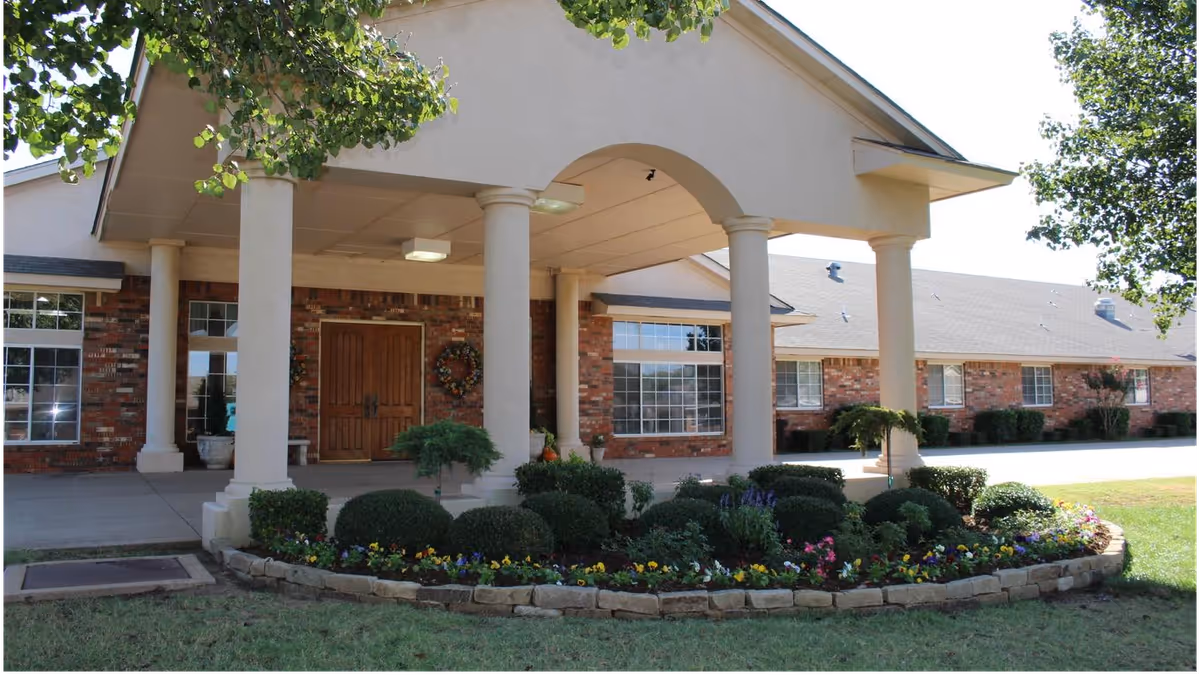Front entrance of a brick senior living building with a covered portico supported by columns, double wooden doors, and a landscaped flower bed.