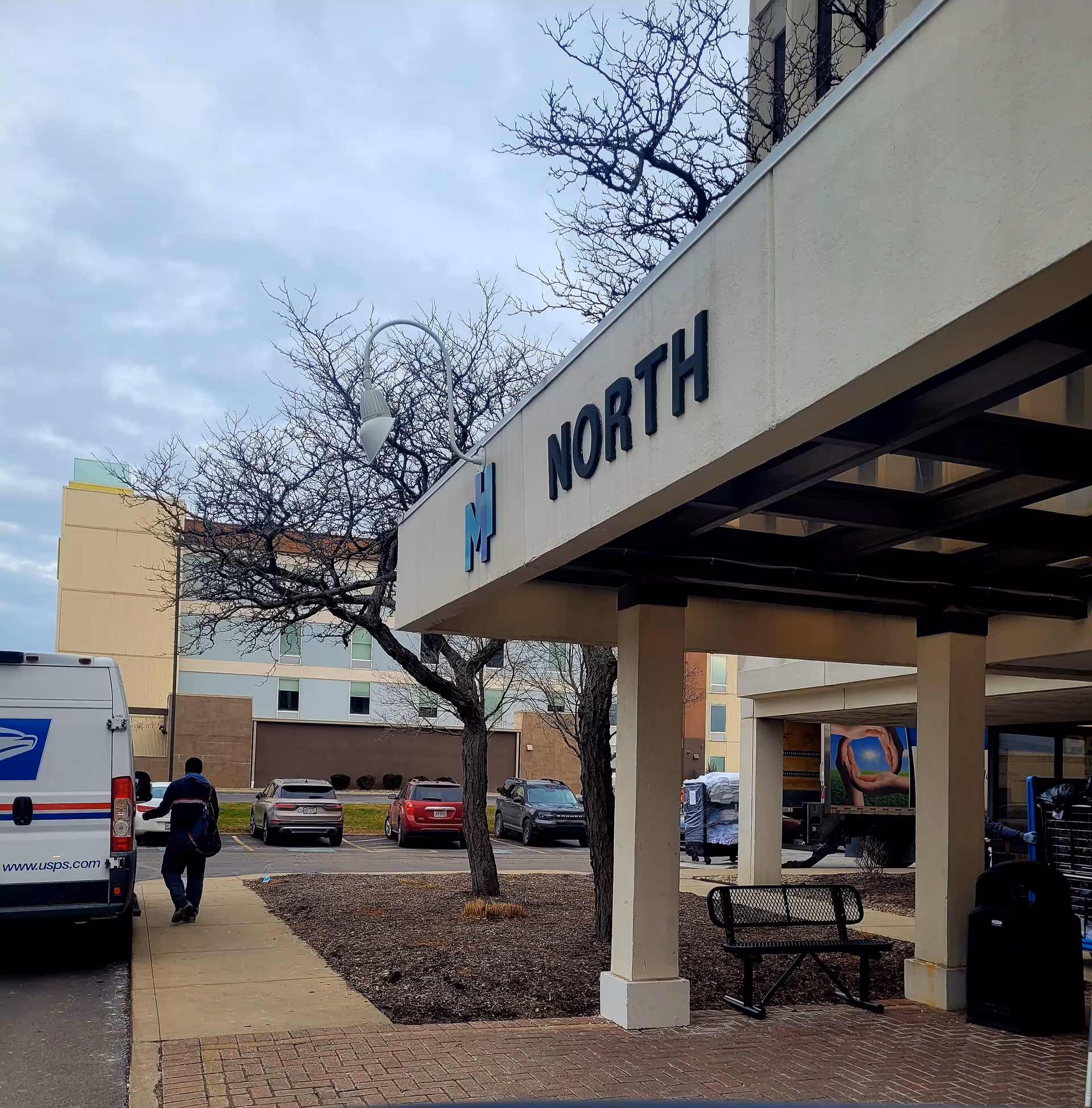 Exterior view of a health center entrance with a bench, trees, and parked cars.