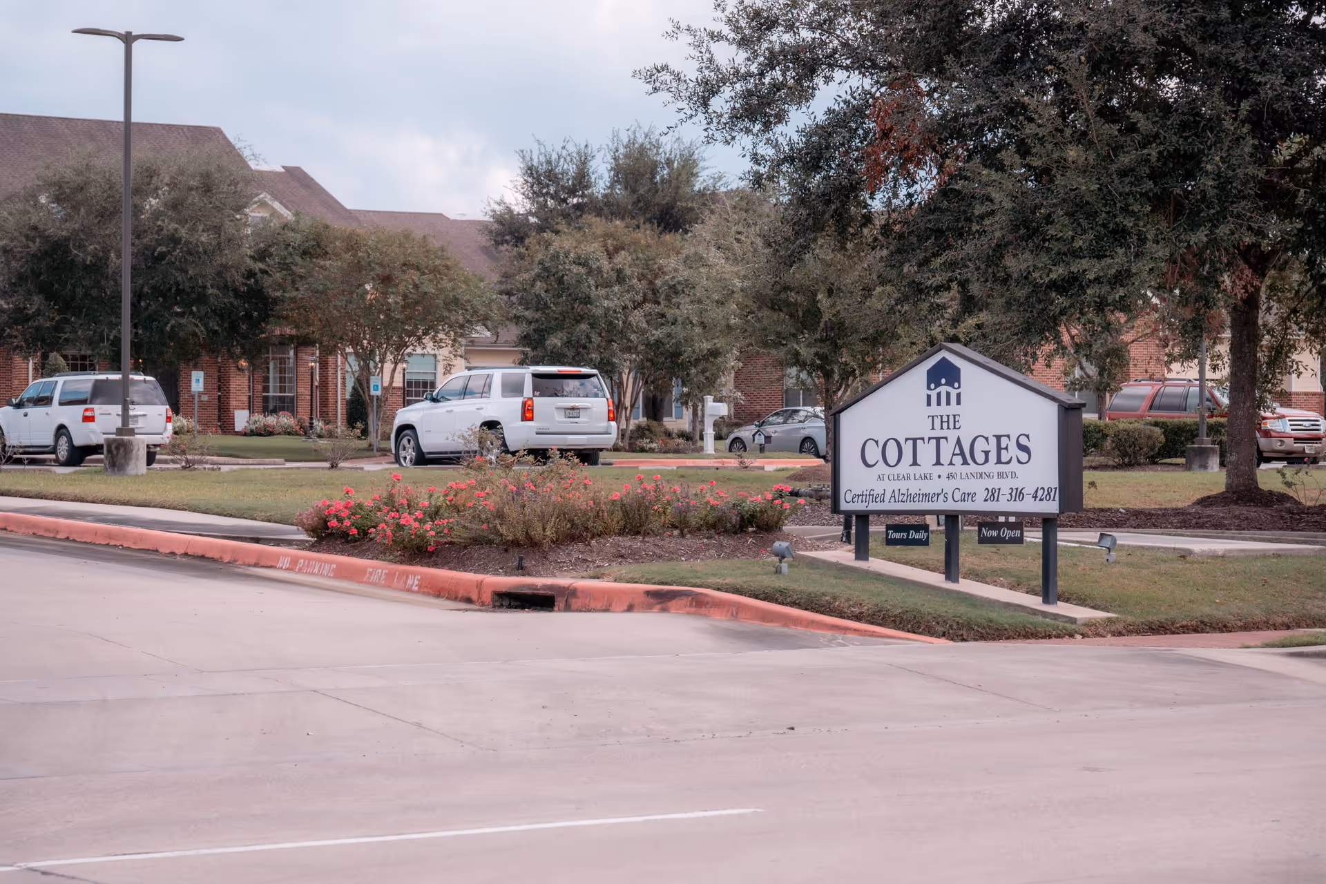 Sign for The Cottages at Clear Lake on a landscaped front lawn with parked cars and brick buildings in the background.
