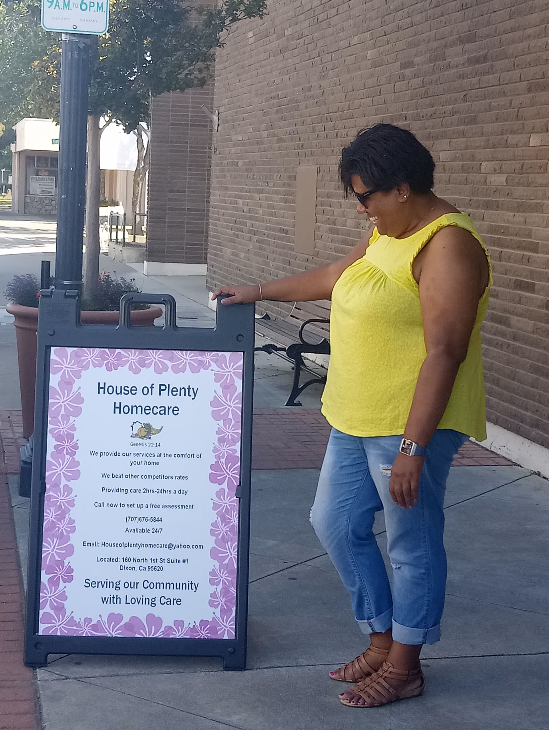 Woman in yellow top stands beside pink bordered House of Plenty Homecare sign outdoors.