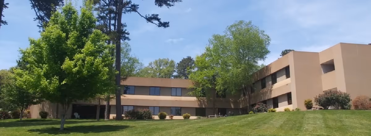 Exterior of The Nursing Center at Little Creek, a beige building with green lawn and trees.