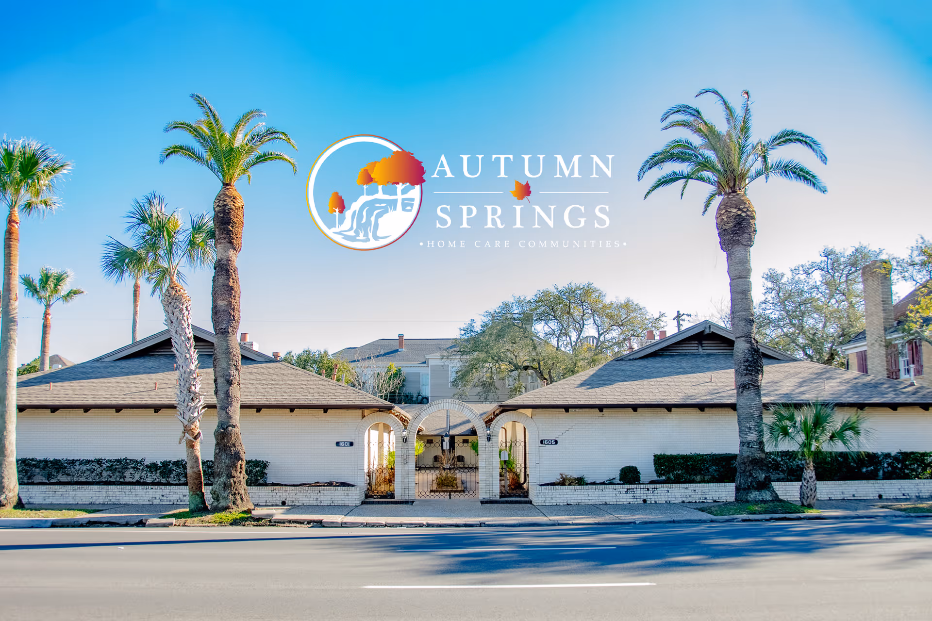 Front exterior view of Autumn Springs Home Care Communities Galveston, showing a white brick building with two peaked roofs, an arched gated entrance in the center, and several tall palm trees in front under a clear blue sky.