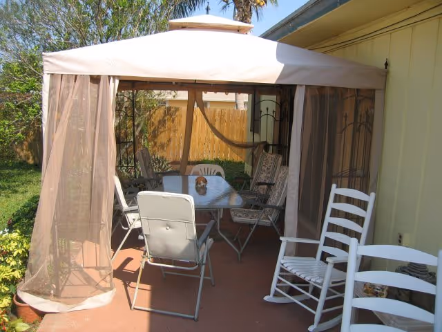Outdoor patio with beige canopy, metal chairs, and a glass table beside a yellow building.