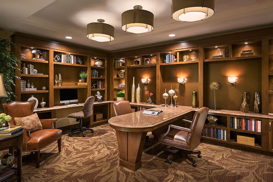 A cozy office or library room with wooden built-in bookshelves filled with books and decorative items. There is a large wooden desk with a marble top surrounded by leather chairs on wheels. A computer is placed on a corner desk area. The room is warmly lit with ceiling lights and wall sconces, and the floor is covered with a patterned carpet.