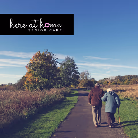 Two elderly individuals walk along a countryside path beside tall grasses under blue sky.