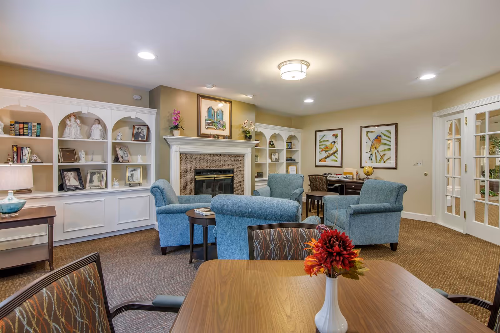 Cozy senior living lounge with blue armchairs, fireplace, built-in shelves, and a round table foreground.
