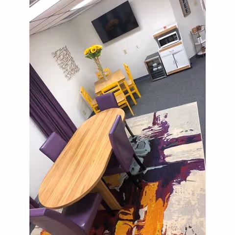 A dining area with a wooden table, purple chairs, flowers, and a kitchenette.