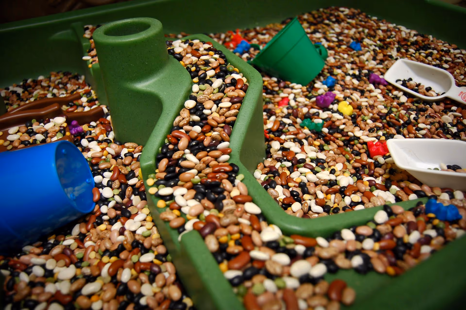 Colorful pebble-filled play area with a green plastic track, blue cup, and scattered bowls.