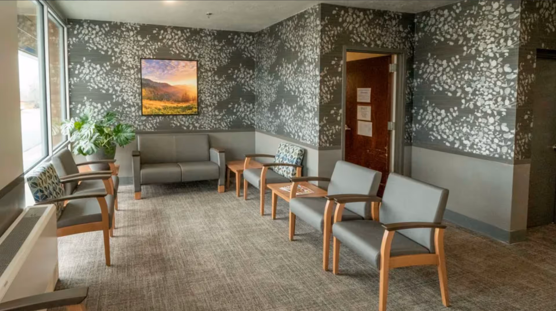A waiting area with several gray chairs with wooden armrests arranged along the walls. The walls have a dark gray and white leaf pattern wallpaper on the upper half and solid gray on the lower half. There is a large window on the left side letting in natural light, a potted plant near the window, and a framed landscape picture on the far wall. A wooden door with papers posted on it is visible on the right side.