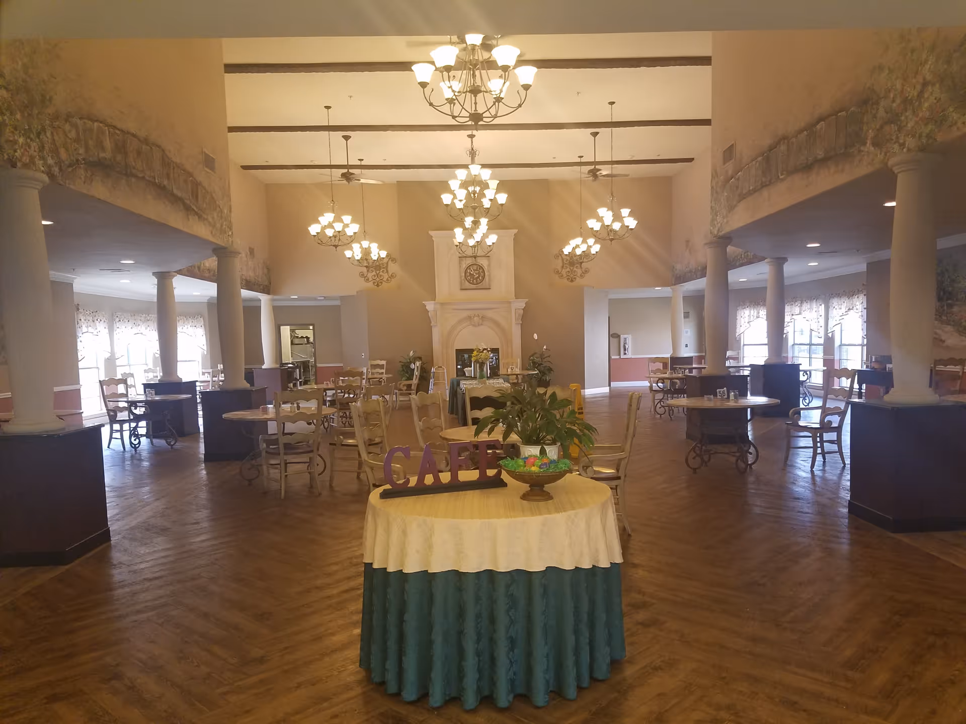Spacious senior living dining hall with chandeliers, columns, and round tables arranged around a central display.