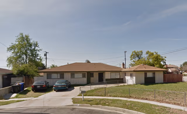 Single-story ranch-style building with a fenced front yard and two cars parked in the driveway under a clear sky.