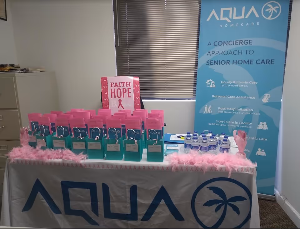 Indoor promotional table with aqua care bags, pink decorations, and a blue Aqua Home Care banner.