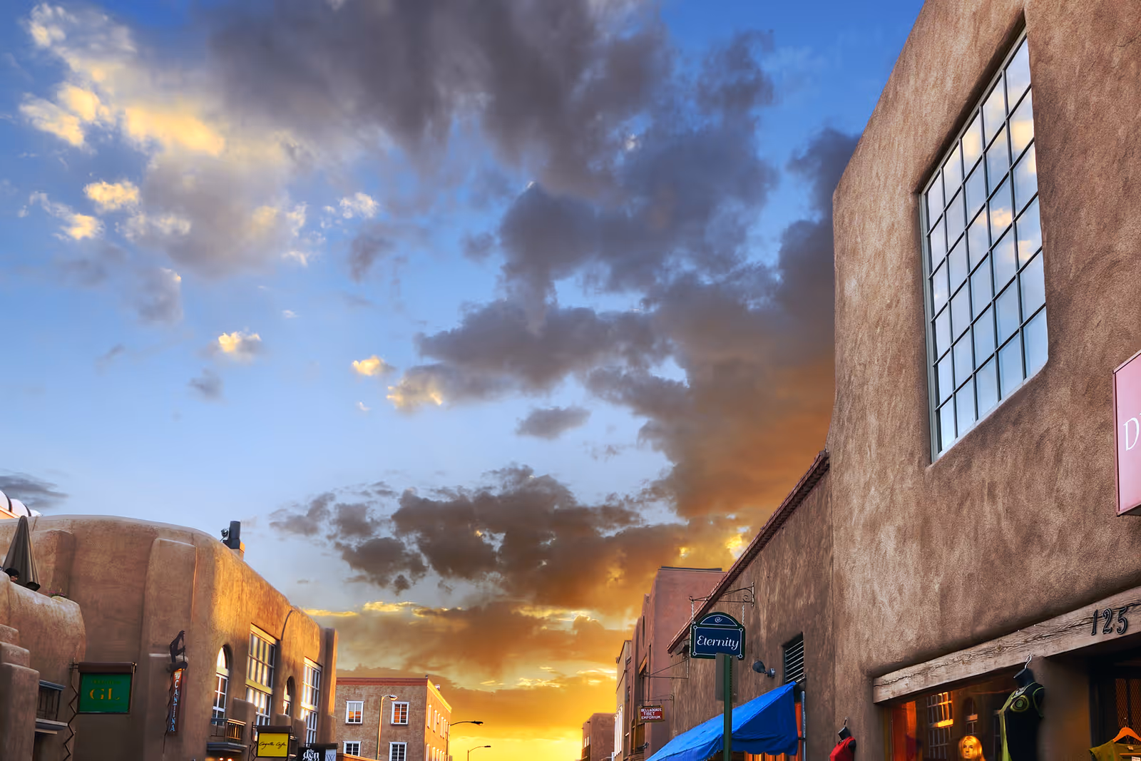 A street view of a southwestern-style building exterior at sunset with a dramatic sky filled with clouds. The buildings have adobe-style architecture with large windows and signs for shops, including one that reads 'Eternity'.