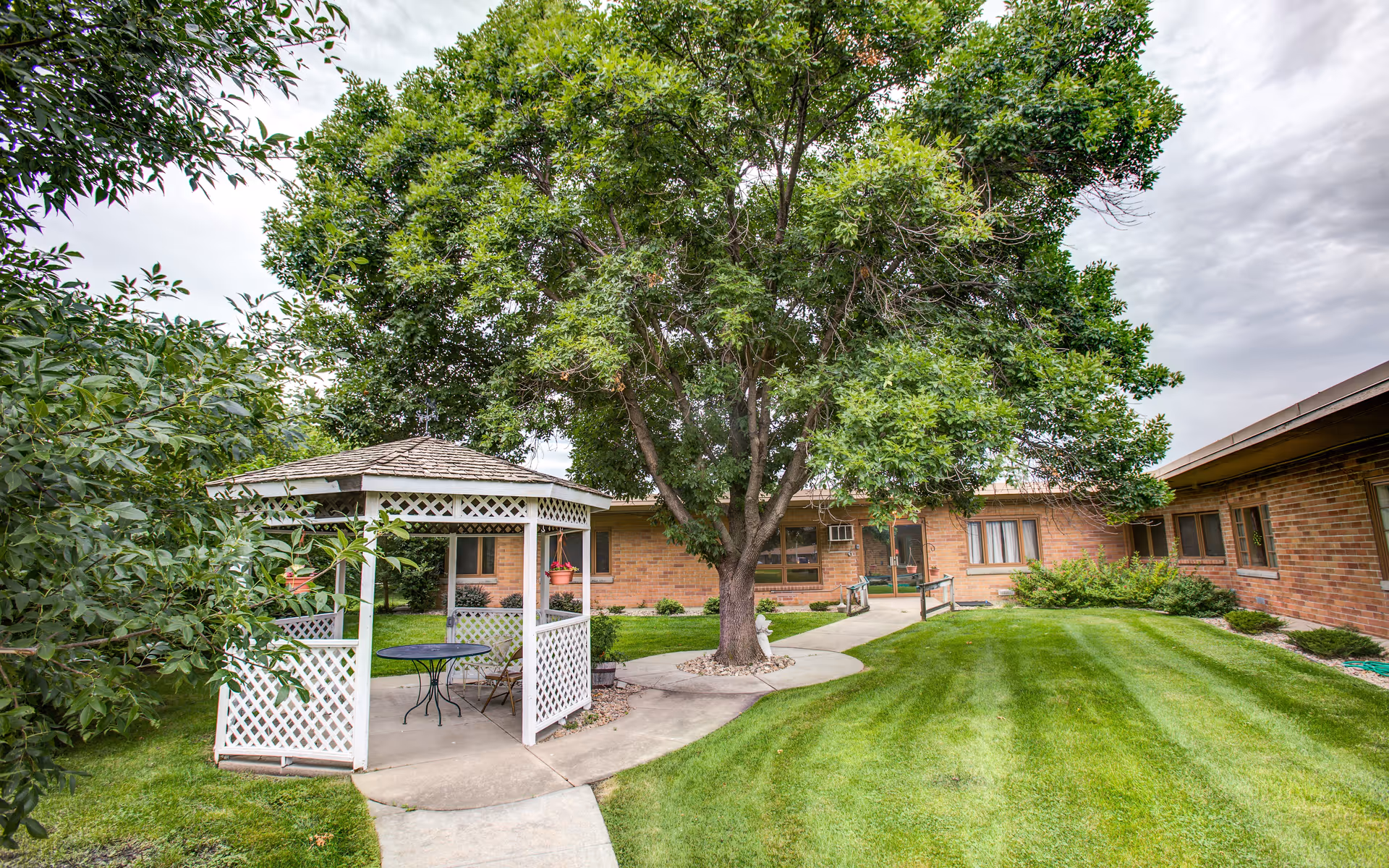 Outdoor garden area at The Cedars at Broadwell featuring a white gazebo with a table and chairs, a large tree in the center, well-maintained green grass, and a brick building in the background under a cloudy sky.