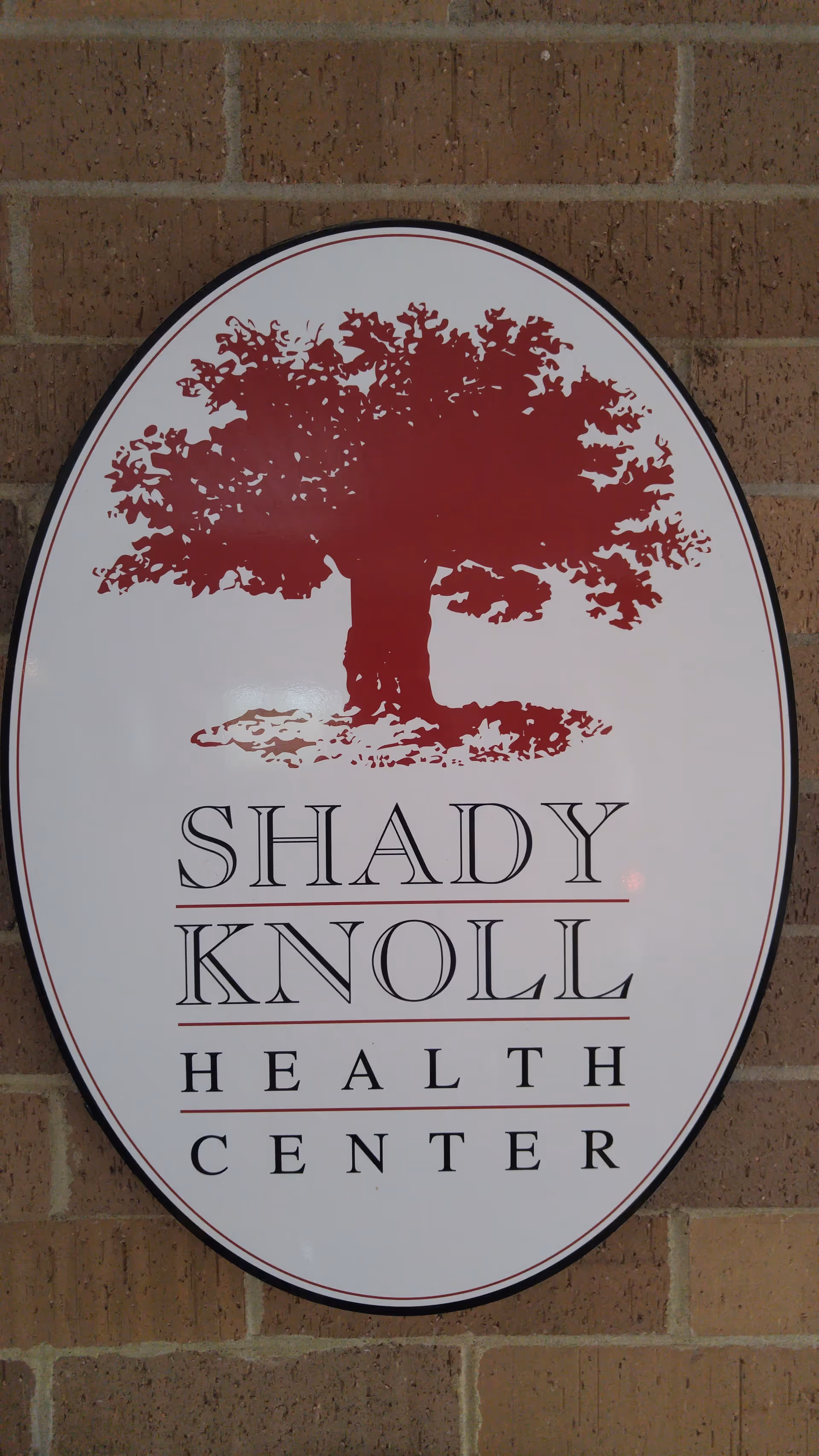 Oval sign with a red tree logo and 'Shady Knoll Health Center' on brick exterior.