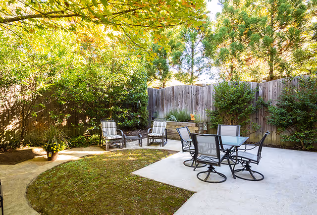 Outdoor patio area with a glass-top table and four chairs on a concrete surface, surrounded by a wooden fence and lush green trees and bushes providing shade and privacy.
