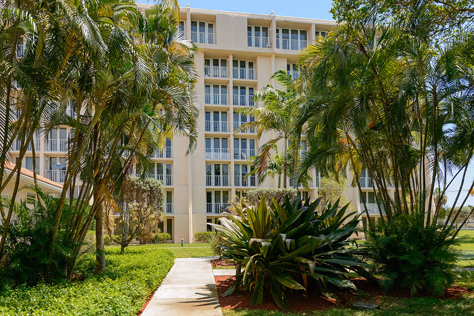 Multi-story residential building with balconies seen from a landscaped courtyard with palm trees and a paved walkway.
