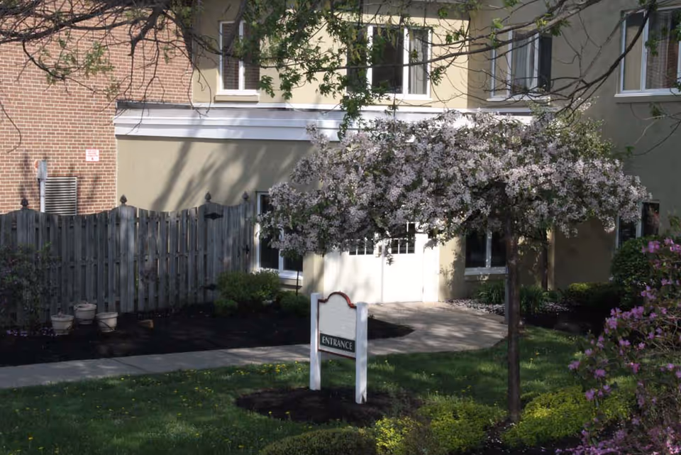 Exterior view of Orchard Brooke Living Center showing a beige building with white-framed windows, a wooden fence, a small tree with white blossoms, green grass, and a sign that reads 'ENTRANCE' near a paved walkway.