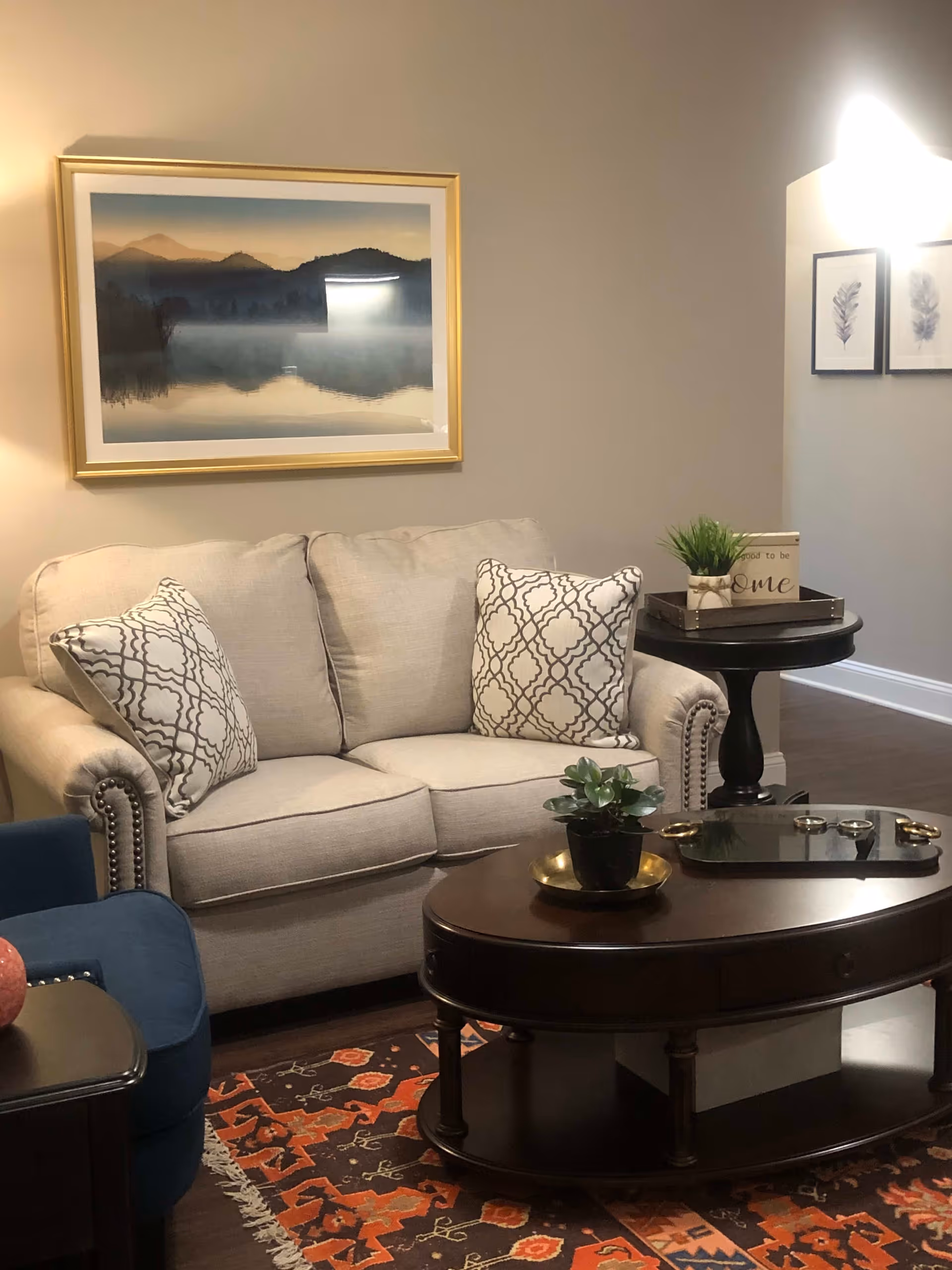A cozy living room area with a beige loveseat adorned with two patterned throw pillows. In front of the loveseat is a dark wooden oval coffee table with a small potted plant and decorative tray. To the left, part of a blue armchair is visible. The floor has a colorful patterned rug. On the wall above the loveseat, there is a framed landscape painting of misty mountains and a lake. To the right, a small round side table holds a potted plant and a decorative sign. The walls are painted a neutral color and the room has warm lighting.