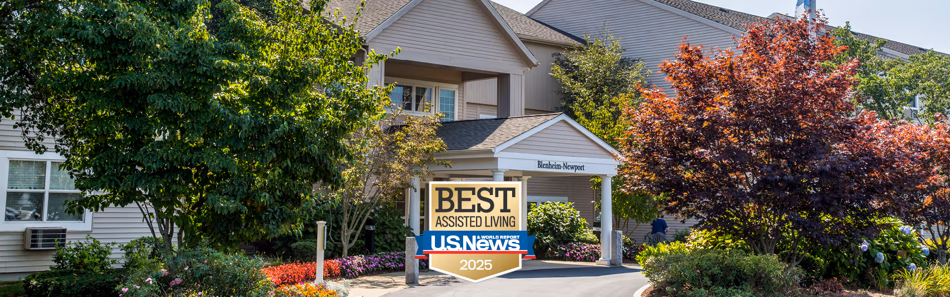 Front entrance of the Blenheim-Newport assisted living building with landscaped trees and a 'Best Assisted Living U.S. News 2025' sign.