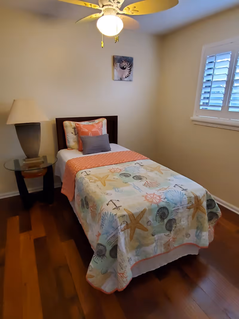A small bedroom with a single bed covered in a seashell and starfish patterned quilt. The bed has three pillows, one coral with white starfish, one with a seashell pattern, and a smaller gray pillow. Next to the bed is a glass-top side table with a large beige lamp. The room has wooden flooring, a ceiling fan with a light, and a window with white shutters.