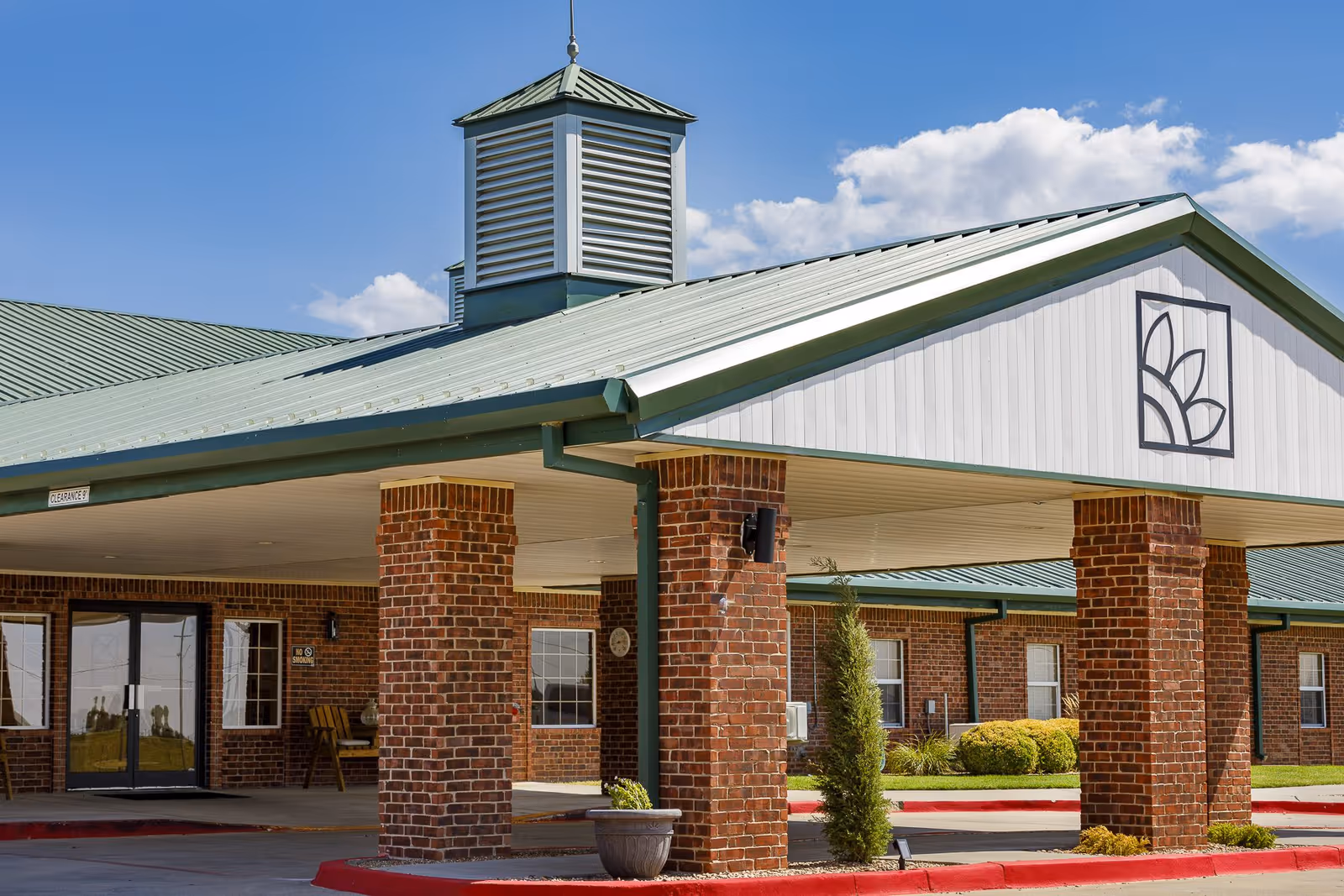 Entrance of a single-story brick assisted living facility with a covered drop-off supported by brick columns and a green metal roof with a logo on the gable.