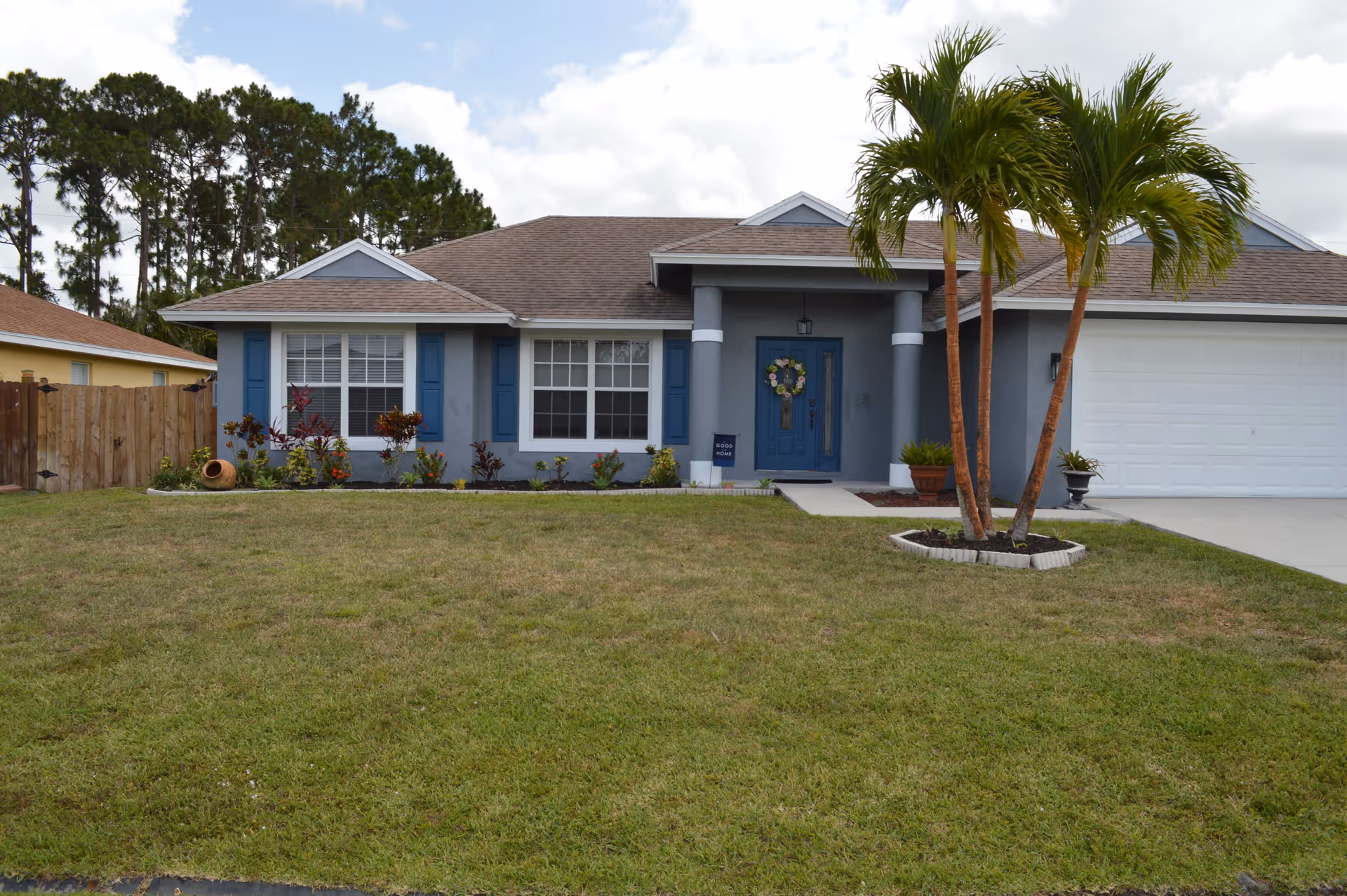 Blue-gray single-story house with front yard, palm trees, and a wreath on the door.