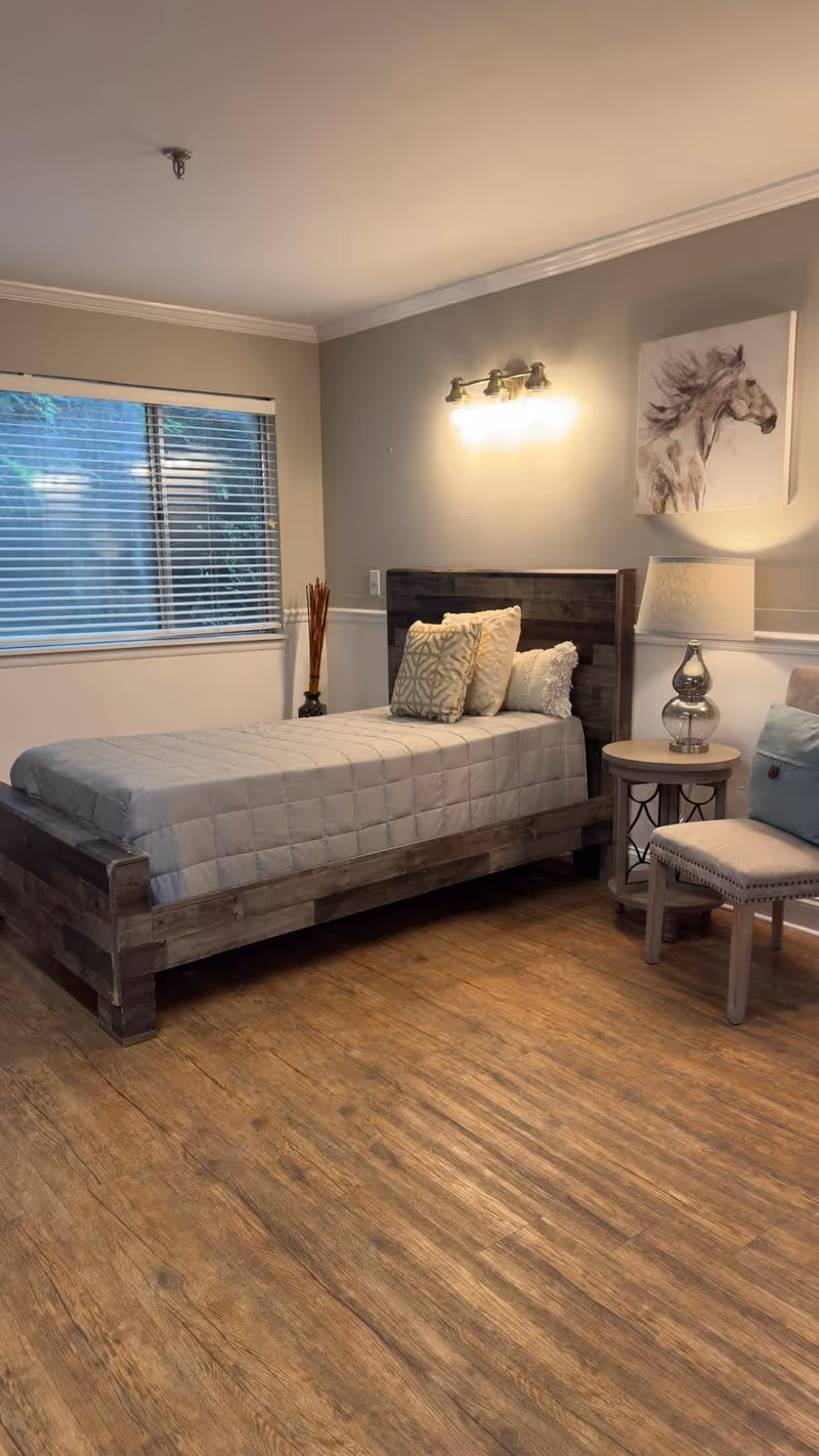 A cozy bedroom with a wooden bed frame and a neatly made bed with a beige quilt and decorative pillows. There is a window with blinds on the left wall, a wall-mounted light fixture above the bed, a painting of a horse on the wall, a round wooden side table with a silver lamp, and a cushioned chair with a blue pillow on the right side. The floor is wooden.