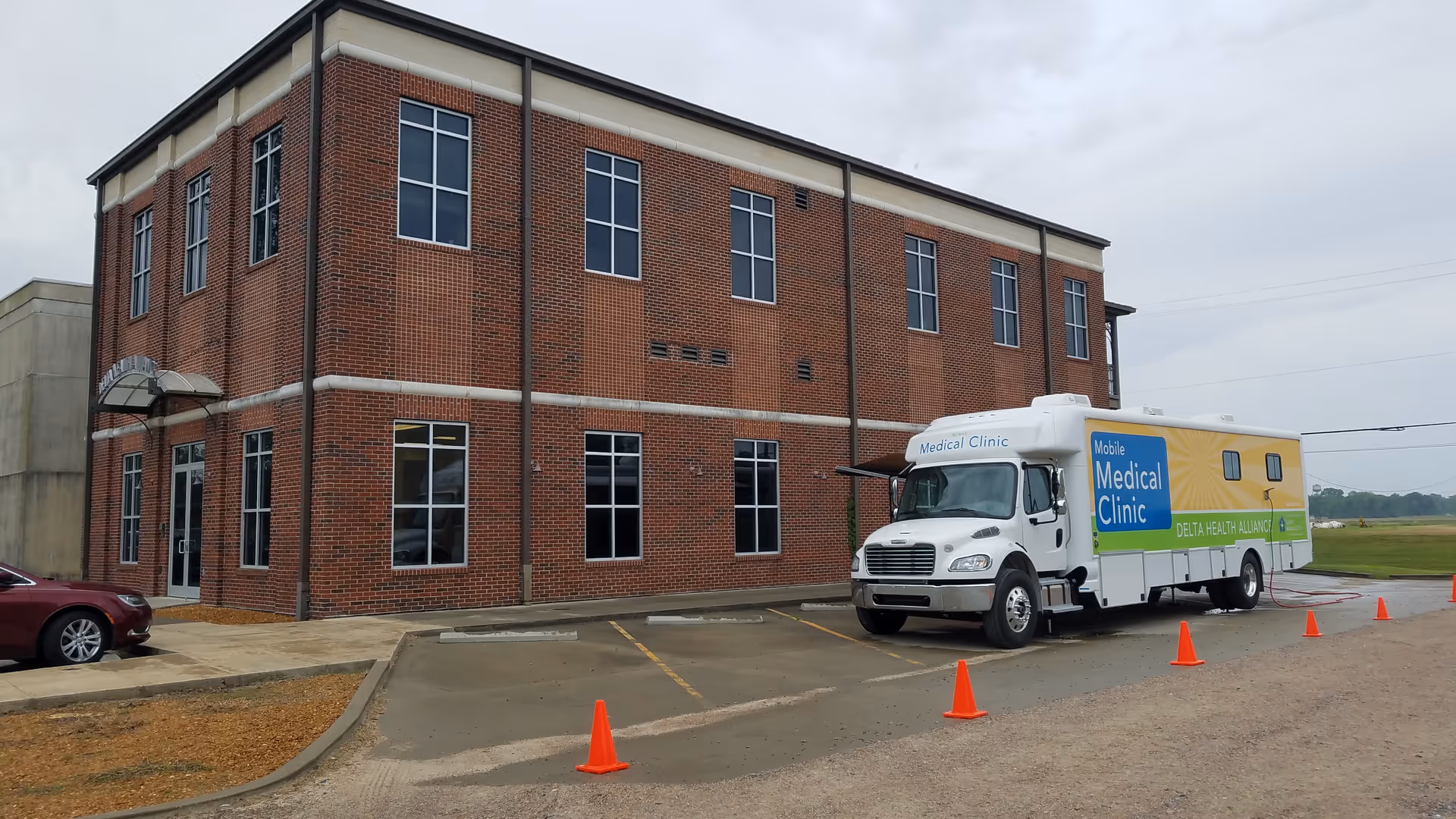 Two-story red brick building beside a mobile medical clinic truck and orange cones.