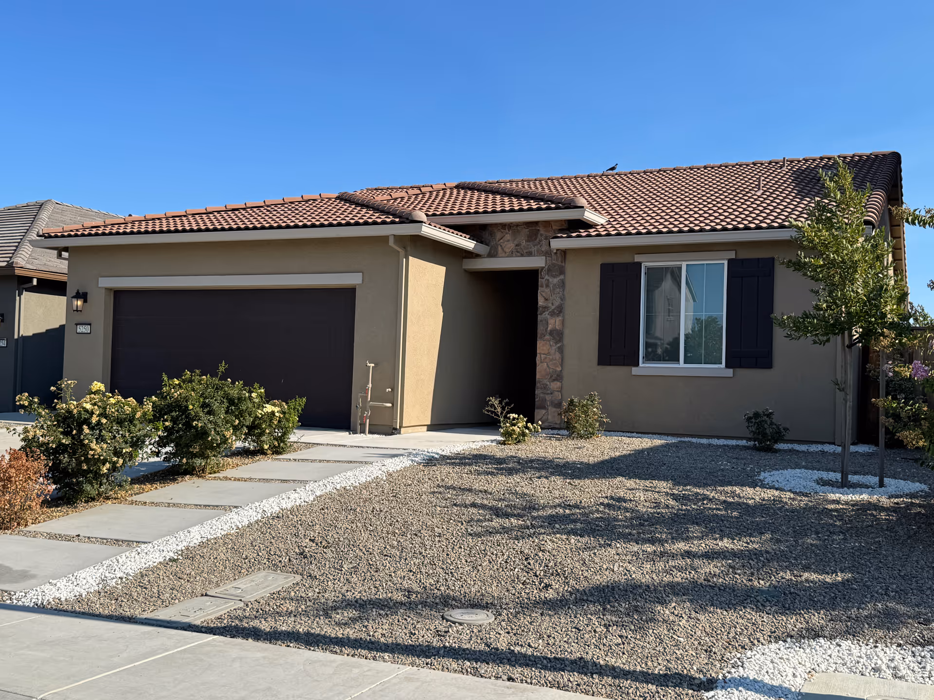 Single-story tan house with brown tile roof, two-car garage, and xeriscaped front yard.