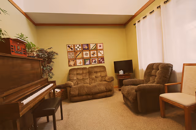 Cozy living room with brown recliners, piano, plants, and quilt wall hanging.