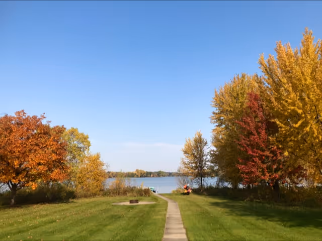 Sunlit autumn park with a paved path along a lake and colorful trees.