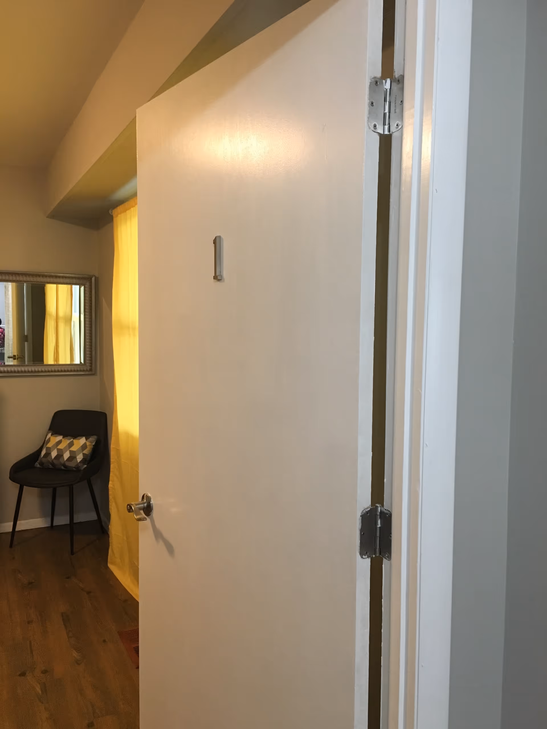 Partially open white door with the number 1 on it, leading into a room with wooden flooring, a black chair with a patterned cushion, a large mirror on the wall, and a yellow curtain covering a window.